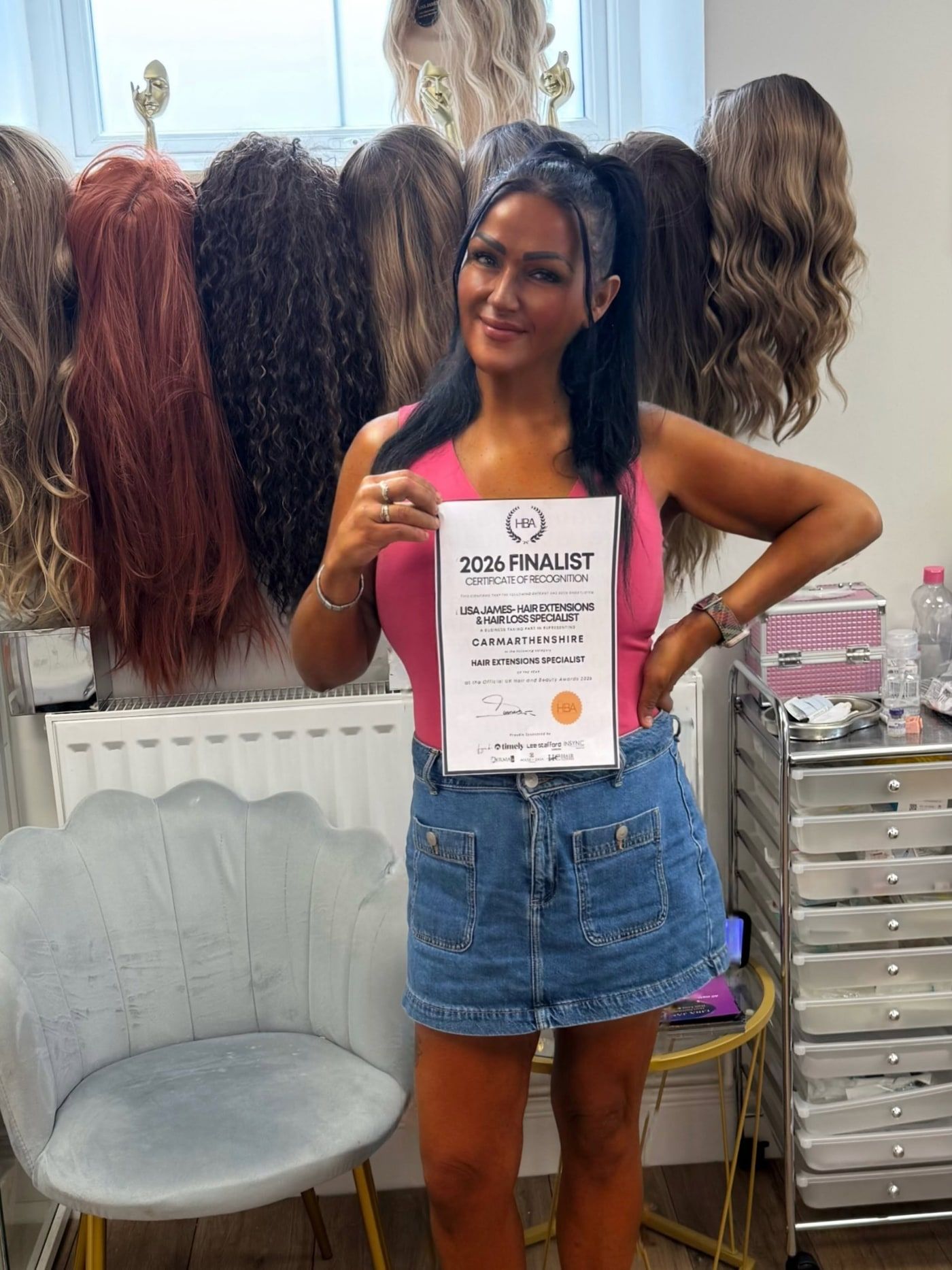 Lisa in her salon holding her 2026 UK Hair & Beauty Awards finalist certificate in front of a wall of wigs